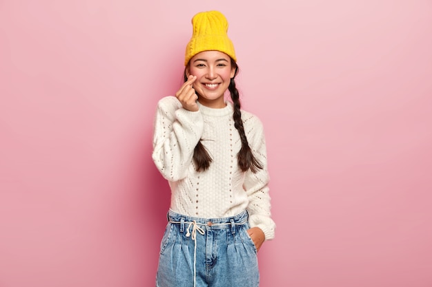 Симпатичная азиатская девушка держит пальцы, сложенные в виде сердечка, показывает корейский знак любви, носит стильную желтую шляпу, белый свитер и джинсы, темные волосы зачесаны в две косички, позирует на фоне розовой стены