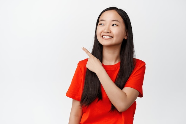 Довольно азиатская девушка улыбается, указывая и глядя в верхний левый угол пустое пространство, показывая логотип или рекламу продажи с довольным выражением лица на белом фоне