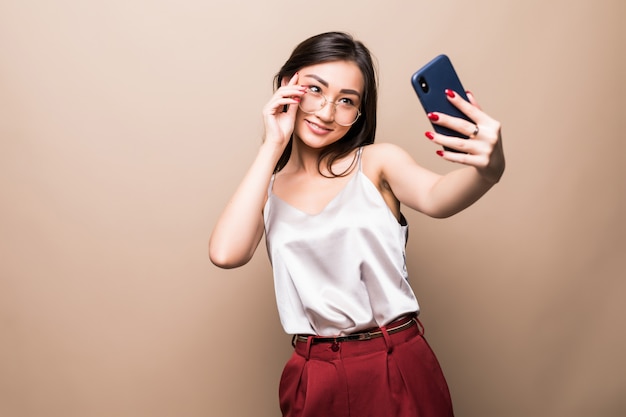 かなりアジアの少女は、ベージュの壁に分離された彼女のスマートフォンでselfieを取る。