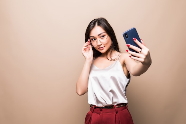 かなりアジアの少女は、ベージュの壁に分離された彼女のスマートフォンでselfieを取る。