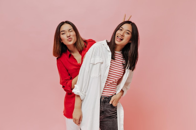 Pretty Asian girls in stylish outfits make funny faces and pose on pink isolated background Woman in red shirt gives bunny ears to her friend