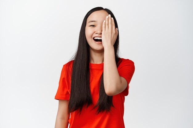 Красивая азиатка закрывает одну сторону лица и беззаботно улыбается, смеясь, стоя на белом фоне в красной футболке