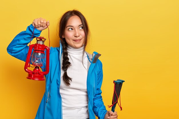 Pretty Asian woman holds oil lamp, looks thoughtfully aside, holds trekking poles and kerosene lamp, has rest in camp near forest poses over yellow wallconcept