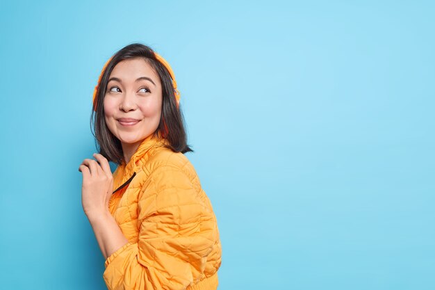 Pretty Asian woman with dark hair stands sideways indoor wears wireless headphones on ears for listening music enjoys good sound quality dressed in orange jacket isolated over blue wall