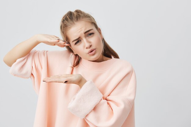 Pretty attractive charming girl wearing stylish pink long-sleeved sweatshirt showing size of something with hands, actively gesturing, frowning her face and posing .