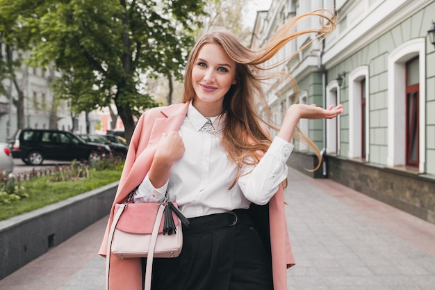 Довольно привлекательная стильная улыбающаяся женщина гуляет по городской улице в розовом пальто, весенняя мода, держит сумочку, элегантный стиль, размахивает длинными волосами
