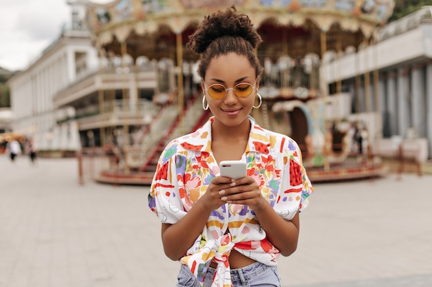 Pretty attractive woman in denim pants, colorful bright shirt and orange sunglasses holding phone and messaging outside