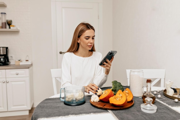 Довольно привлекательная женщина с темными волосами в белой рубашке ищет в смартфоне рецепт Женщина на кухне готовит свежие овощи