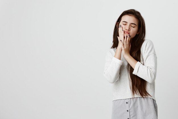 pretty beautiful brunette teenage woman wearing her long dark straight hair loose in casual clothes touching cheeks with hands, posing against gray wall with closed eyes.