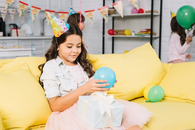 Pretty birthday girl sitting on sofa holding balloon in hand looking at gift box