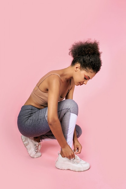 Pretty Black Woman Tying Shoelaces On Pink Background. Wearing Stylish Sportwear.