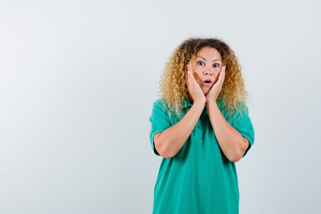 Pretty blond lady in green polo t-shirt keeping hands on cheeks and looking shocked , front view.