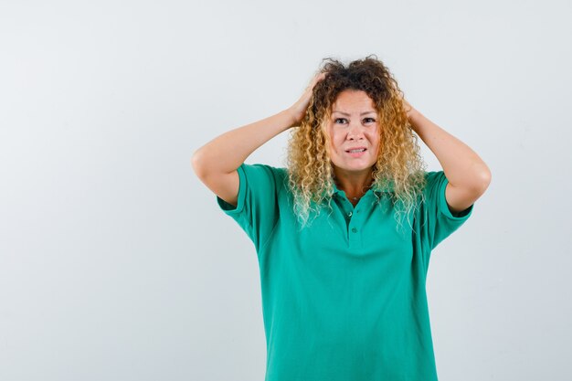 Pretty blond lady keeping hands on head in green polo t-shirt and looking helpless , front view.