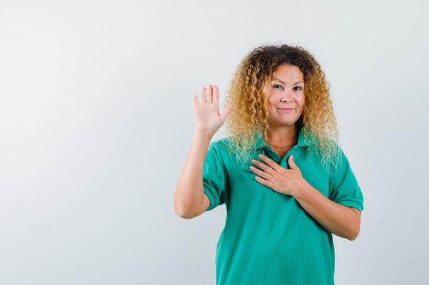 Pretty blond lady showing stop sign, keeping hand on chest in green polo t-shirt and looking confident. front view.