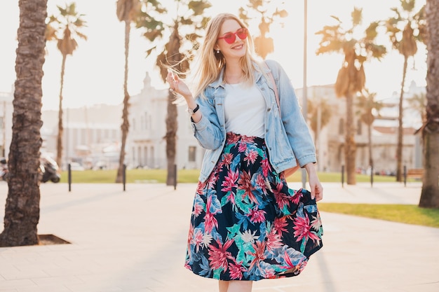 Pretty blond smiling woman walking in city street in stylish printed skirt and denim oversize jacket wearing pink sunglasses