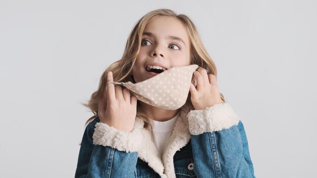 Pretty blond teenager girl taking off protective mask looking happy on camera over white background Safety first concept