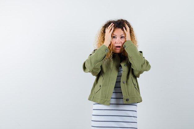 Pretty blond woman keeping hands on head in green jacket and looking perplexed. front view.