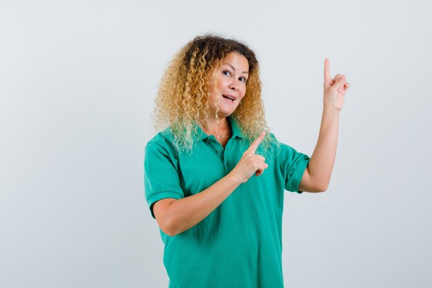 Pretty blond woman pointing up in green polo t-shirt and looking astonished , front view.