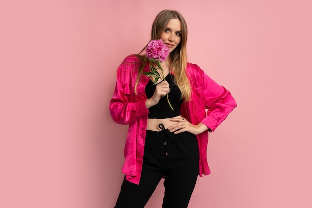 Free photo pretty blond woman posing with peony flower in stylish summer outfit over pink wall