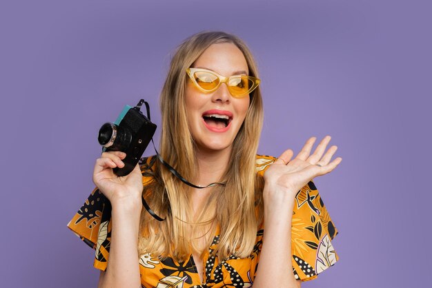 Pretty blond woman in stylish summer outfit holding camera and posing on purple background in studio Travel and summer fashion concept