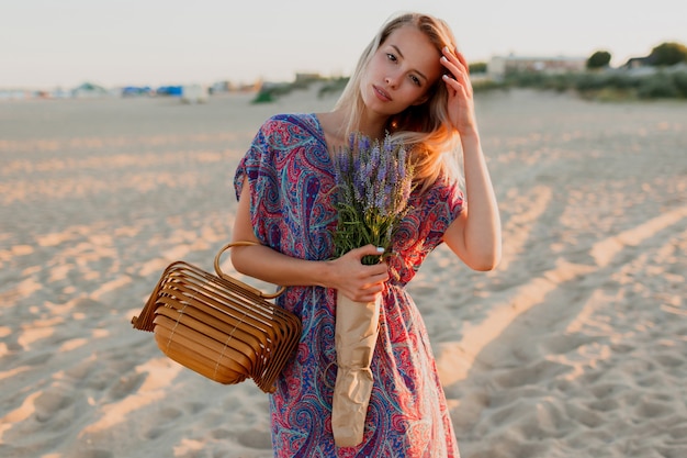 Pretty blond woman with bouquet of lavender walking on the beach. Sunset colors.