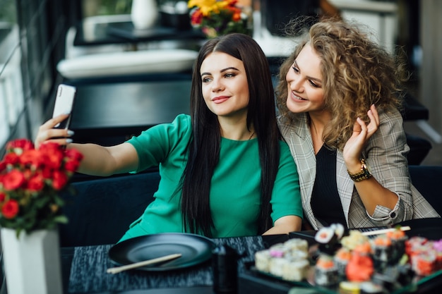 Pretty blonde and brunette girl taking a picture by the cell phone with sushi plate on table. Chenese eat, friends time.