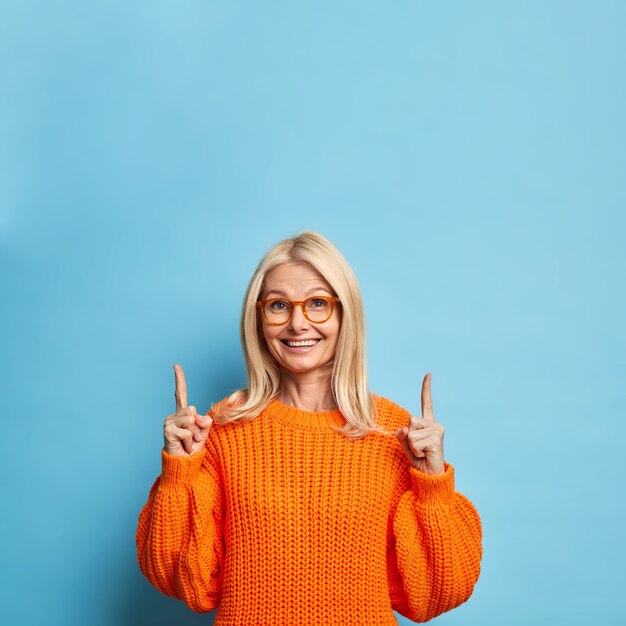 Pretty blonde forty years old woman smiles happily pointing up at copy space, wears spectacles orange sweater.