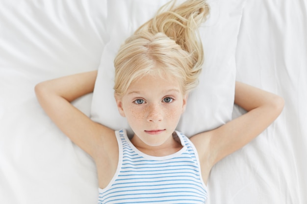 無料写真 彼女の穏やかな輝く目で直接見て、かなり金髪のそばかすのある青い目の女の子が白い寝具のベッドで横になっています。