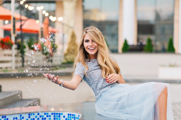 Pretty blonde girl in blue tulle skirt having fun on terrace background. She splashing water and smiling to camera.