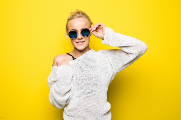 Довольно блондинка в современном белом свитере в блестящих синих солнцезащитных очках позирует на желтом