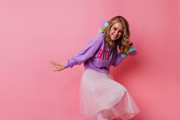 Pretty blonde girl posing with sincere smile on pastel background Pleasant young woman with skateboard dancing and laughing