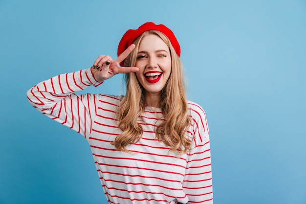 Bella ragazza bionda in camicia a righe che mostra il segno di pace. vista frontale di ridere signora francese in posa sulla parete blu.