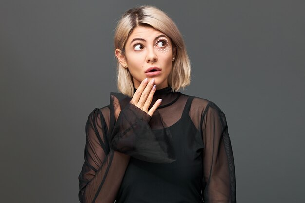 Pretty blonde girl in stylish transparent dress holding hand at her face, looking up with mouth wide opened, canât believe her eyes, feeling amused and astonished. Surprise, shock and astonishment