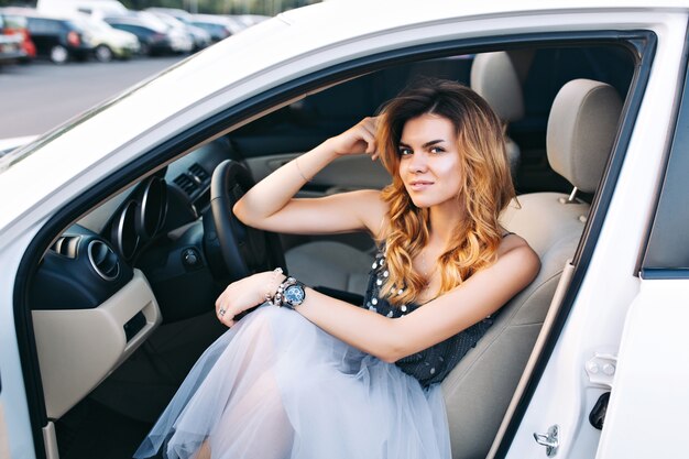 Pretty blonde girl in tulle skirt is sitting in car on parking. She is smiling .