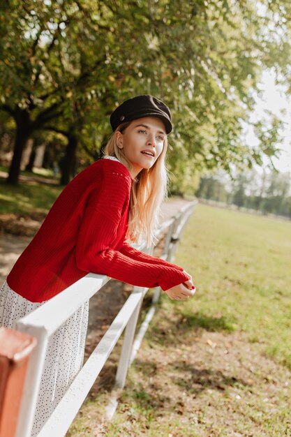 公園のフェンスの近くでポーズをとって自然なメイクのかわいいブロンドの女の子。びっくりした素敵なアクセサリーの美しい女性。