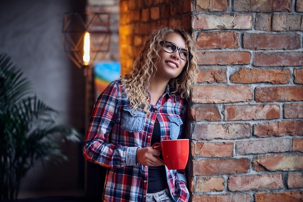 Pretty blonde hipster female drinks morning coffee in a restroom with loft interior.