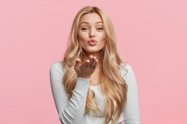 Pretty blonde woman blows passionate kiss expresses her love, isolated over pink wall. Adorable young female model makes air kiss. People, body language and beauty concept