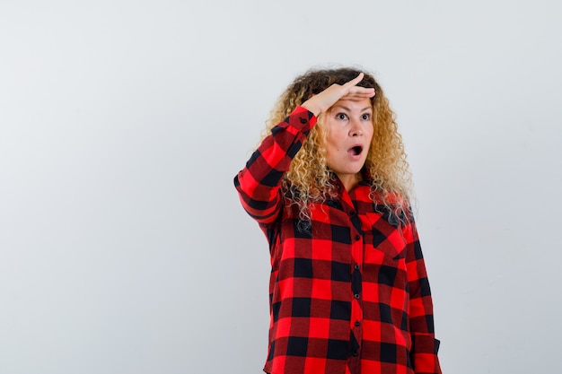 Pretty blonde woman looking far away with hand over head in checked shirt and looking wondered. front view.