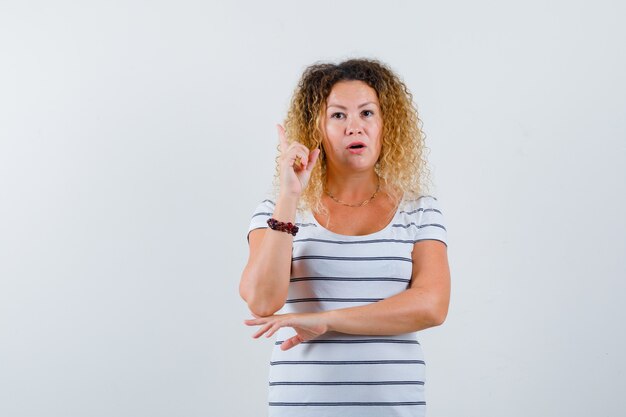 Pretty blonde woman pointing up, showing eureka gesture in striped t-shirt and looking smart. front view.