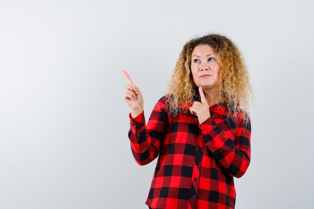 Pretty blonde woman pointing at upper left corner in checked shirt and looking indecisive. front view.