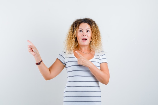 Free photo pretty blonde woman pointing at upper left corner in striped t-shirt and looking surprised , front view.