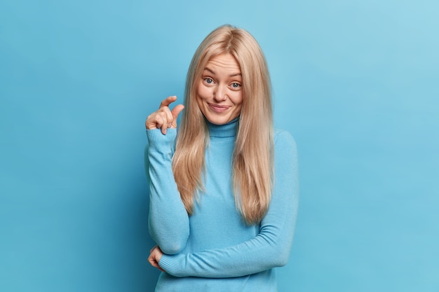 Pretty blonde woman shapes small invisible item with fingers demonstrates small amount of something talks about her little achievement dressed in casual turtleneck 