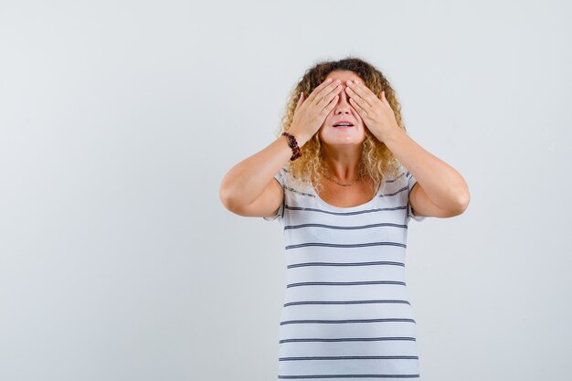 Pretty blonde woman in striped t-shirt covering eyes with hands and looking excited , front view.