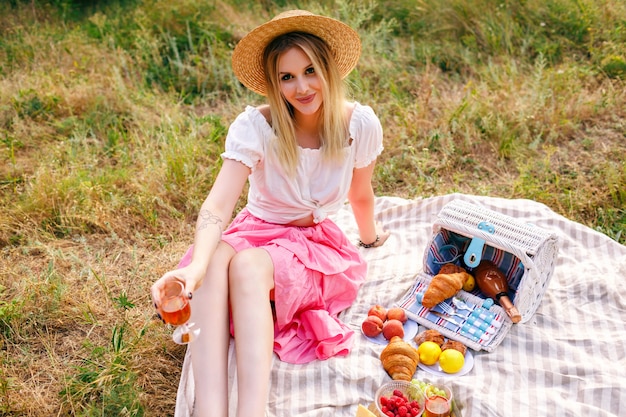 Pretty blonde woman wearing vintage style outfit, enjoying countryside picnic in French style, drinking wine with croissants and fruits