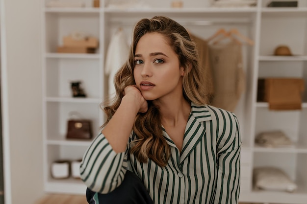 Pretty blonde woman in white and green striped silk shirt leans on her knees and looks into camera