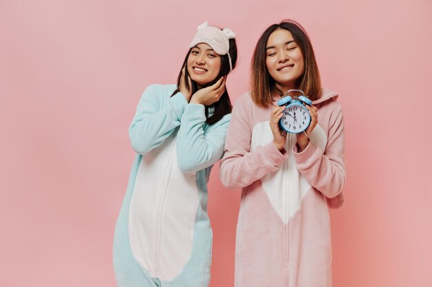 Pretty brunette Asian girls in sleep wears smile sincerely Young women in white pajamas pose on pink background Woman in kigurumi holds alarm clock