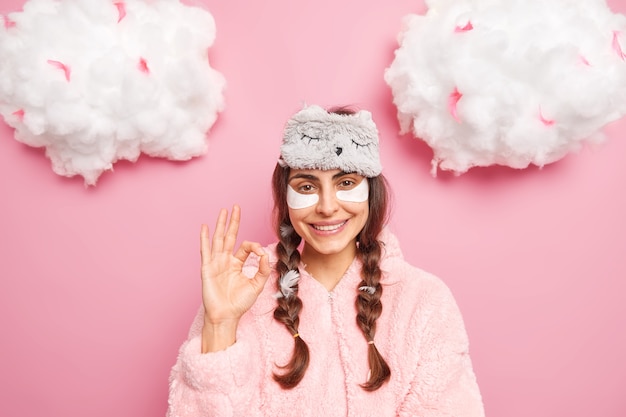 Pretty brunette Caucasian woman smiles gently shows okay gesture likes something applies collagen patches to reduce puffiness under eyes dressed in soft nightwear isolated over pink wall