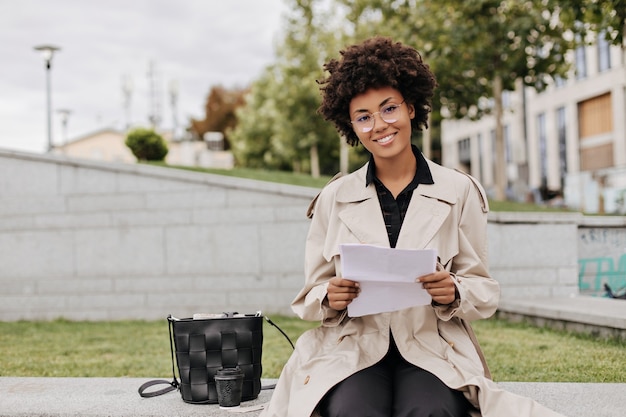 Pretty brunette curly woman in eyeglasses and beige trench coat holds paper sheet, smiles and sits outdoors