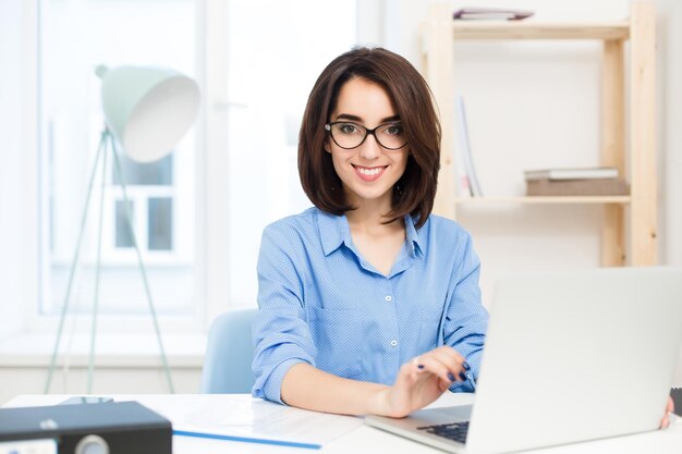 파란색 셔츠를 입은 예쁜 갈색 머리 소녀가 사무실의 테이블에 앉아 있습니다. 그녀는 노트북으로 작업하고 카메라를 향해 웃고 있습니다.
