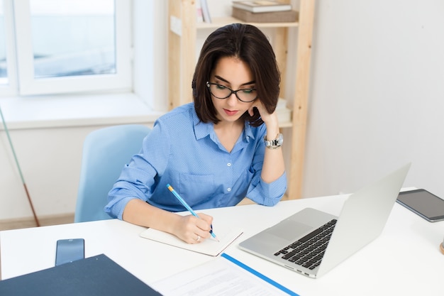 A pretty brunette girl in a blue shirt  is working at the table in office.  She is writing in notebook seriously.